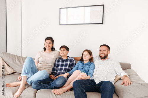 Image of caucasian family with two children resting in living room at home, and looking at tv together while sitting on sofa