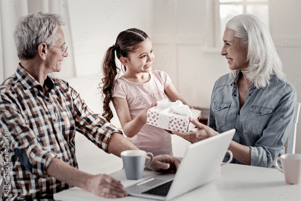 Love you granny. Positive pleased grandma having gift from little granddaughter while grandpa admiring then sitting in front of laptop Love you granny. Positive pleased grandma having gift from little granddaughter while grandpa admiring then sitting in front of laptop