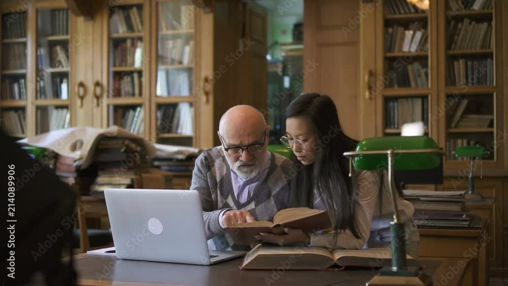Academic lecturer and female student discussing theme at table in university library, people are talking, looking in book at desk with laptop on background of bookcases during educational day. Concept