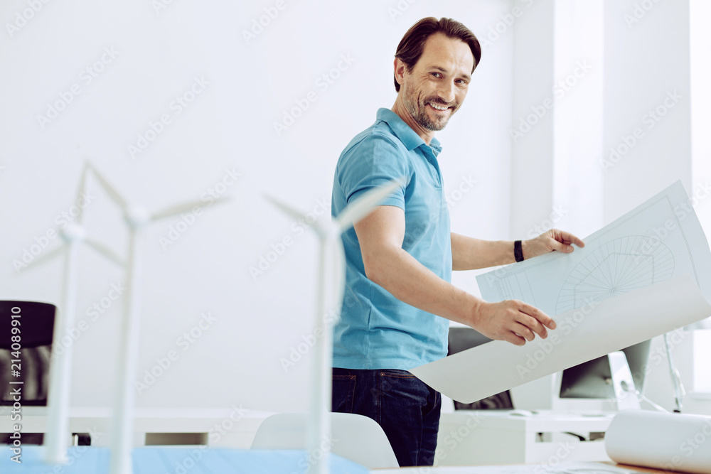 Perfect model. Smiling positive skilled engineer looking at the perfect models of windmill generators while holding a big drawing in his hands