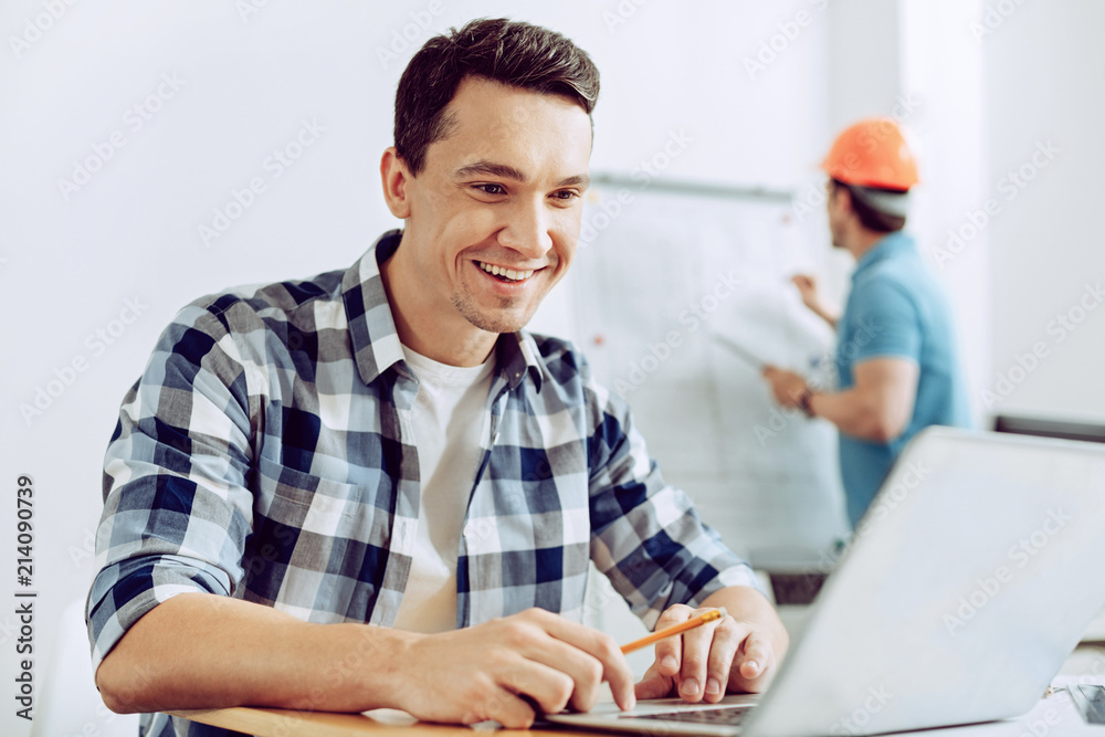 Happy employee. Smiling cheerful positive young worker looking at the screen of a laptop while being in his office