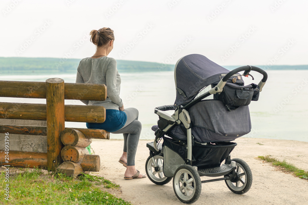 Naklejka premium Young sporty mother with stroller sitting on wooden bench near lake or river. Mom walking with baby in pram near pond at early morning. Healthy outdoor sleeping. Toned