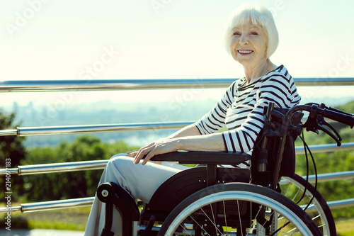 Joyful mood. Cheerful mature woman sitting on wheelchair and keeping smile on her face while breathing fresh air
