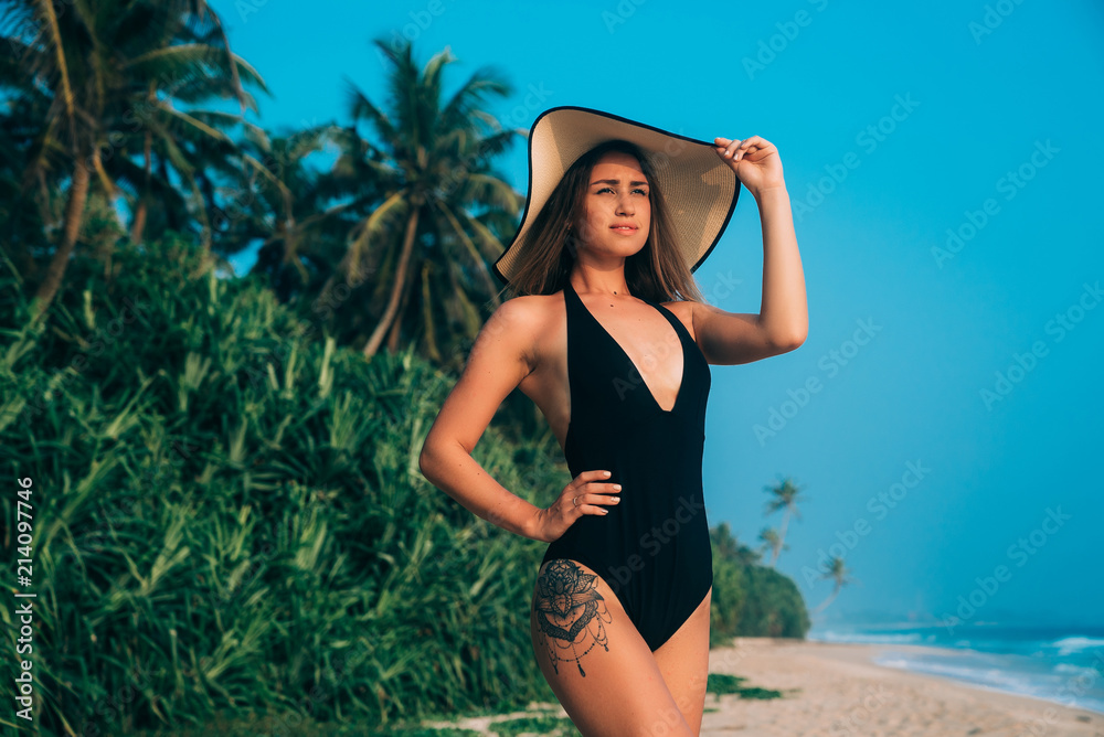 Angle from below, a young girl in a swimsuit on the beach admires the horizon, enjoys relaxation in her expensive hotel, wears a stylish hat, against the blue sky and green jungle Angle from below, a young girl in a swimsuit on the beach admires the horizon, enjoys relaxation in her expensive hotel, wears a stylish hat, against the blue sky and green jungle