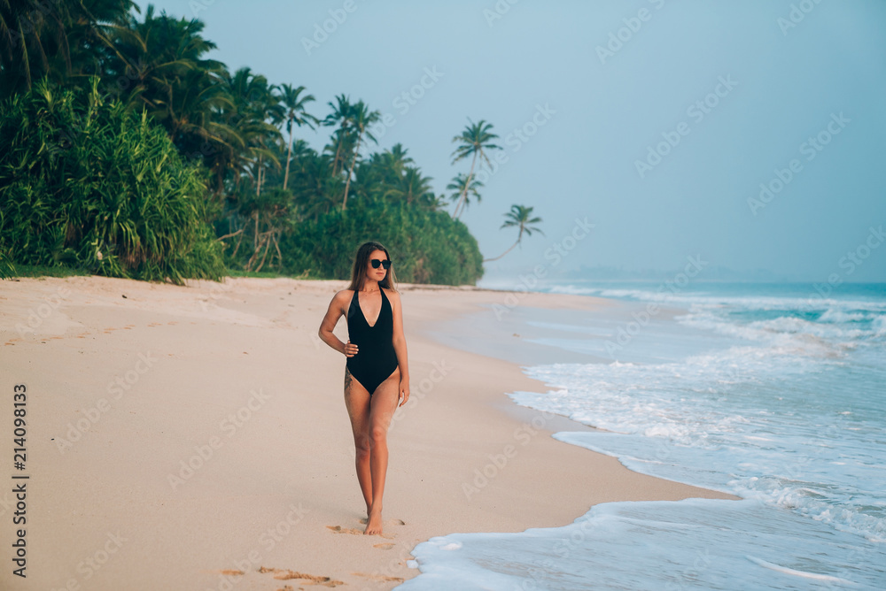 A stylish European strolls along the seashore, holds her hand on her thin waist, wears trendy sun glasses