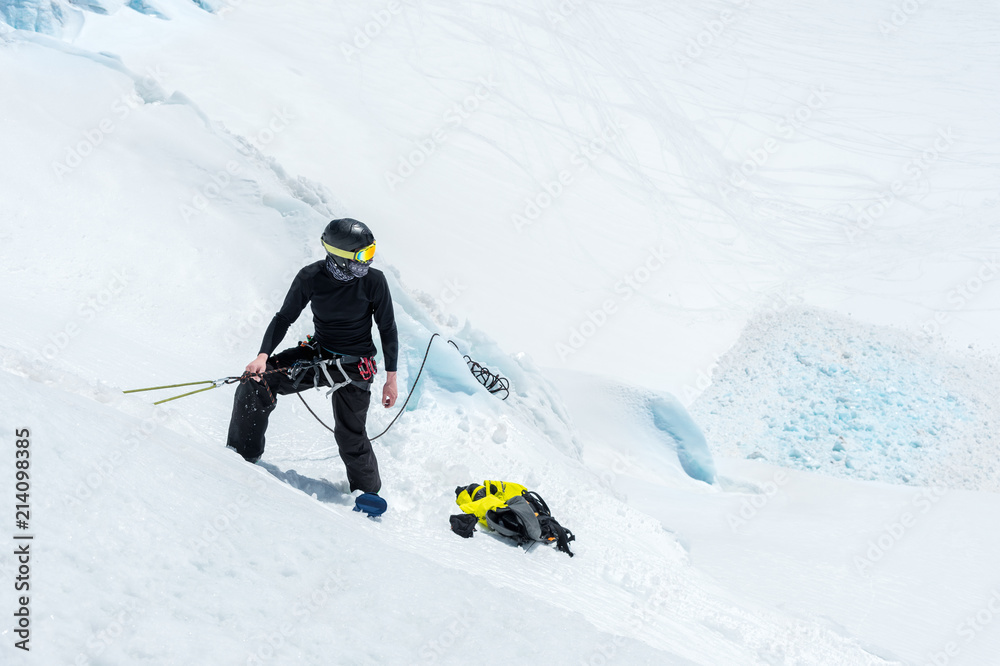 A professional mountaineer in a helmet and ski mask on the insurance does notch the ice ax in the glacier. The work of a professional climber in winter on a glacier against the blue sky A professional mountaineer in a helmet and ski mask on the insurance does notch the ice ax in the glacier. The work of a professional climber in winter on a glacier against the blue sky