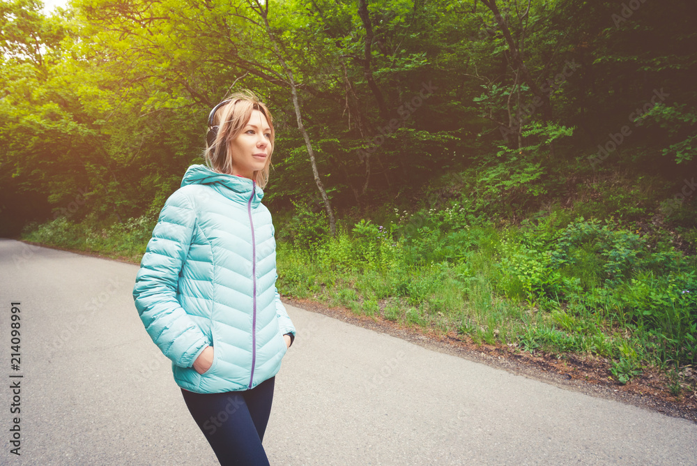 Portrait of an attractive sports girl blonde in a light running down jacket dressing bluetooth headphones with music or the sounds of nature while on a forest road Portrait of an attractive sports girl blonde in a light running down jacket dressing bluetooth headphones with music or the sounds of nature while on a forest road