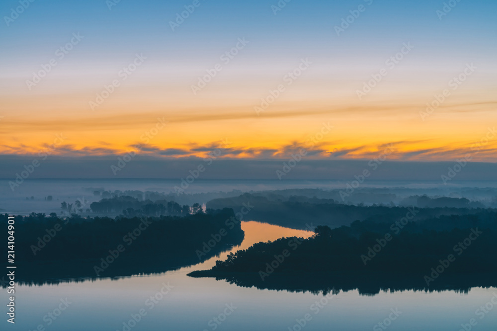 Early blue sky reflected in river water. Riverbank with forest under predawn sky. Yellow stripe in picturesque sky. Fog hid trees on island. Colorful morning atmospheric landscape of majestic nature.