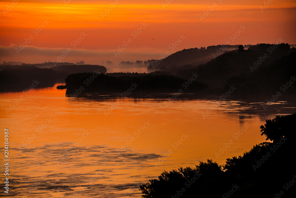 Red dawn sky reflected on water in valley of river. Morning haze in distance above forest on shore. Birds flying in sky at sunrise. Fog on riverbank. Colorful atmospheric landscape of majestic nature.