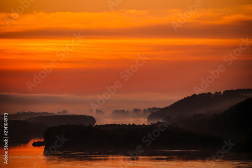 Fototapeta Naklejka Na Ścianę i Meble -  Red dawn sky reflected on water in valley of river. Morning haze in distance above forest on shore. Birds flying in sky at sunrise. Fog on riverbank. Colorful atmospheric landscape of majestic nature.