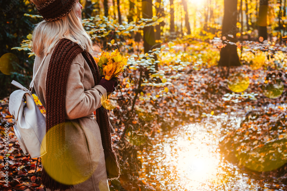 Side view of female holding bouquet of yellow autumn maple leaves in her gloved hands. Ground covered with orange leaves lightened by warm evening backlit sun light reflected in a pond