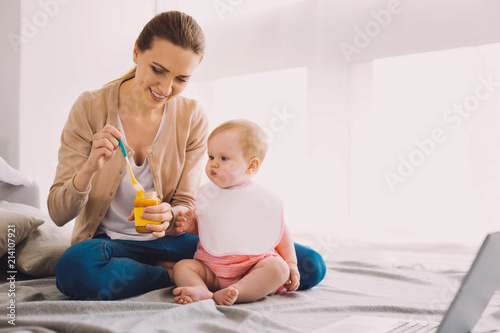 New food. Cheerful attentive babysitter smiling while giving tasty fresh baby food to a cute toddler