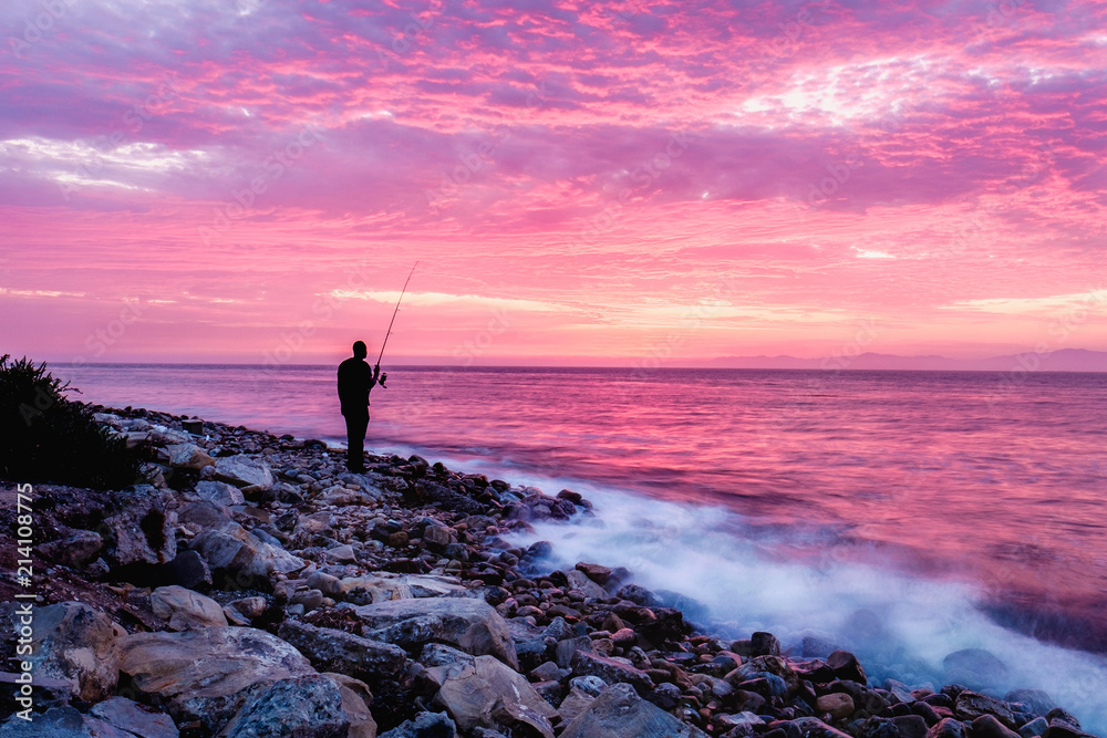 fisherman on rocks on ocean shoreline with colorful sunset skies
