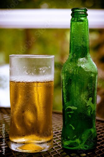 A full glass of beer next to an empty bottle against a blurred nature background.