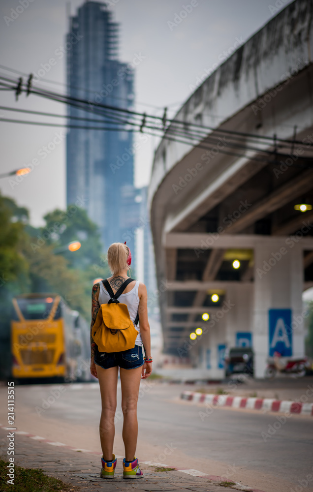 A young girl with tattoos stands on a city street on a background of tall buildings and a bridge. She wears denim shorts and an orange backpack. Holidays in Pattaya A young girl with tattoos stands on a city street on a background of tall buildings and a bridge. She wears denim shorts and an orange backpack. Holidays in Pattaya