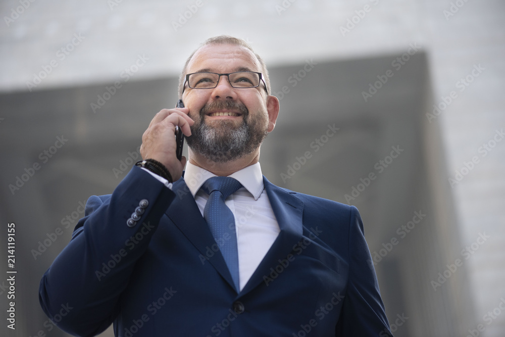 Closeup view of white middle age, bearded corporate businessman wearing a suit and tie with glasses, smiling  walking while talking on the phone