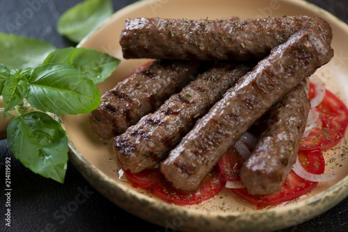 Closeup of grilled balkan cevapi or cevapcici sausages with tomatoes, onion and green basil leaves, selective focus