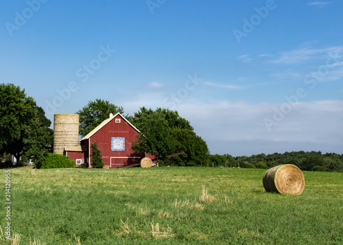 Bright red barn with barn quilt and silo against a bright blue summer sky. Large round hay bales are in the field in front of the barn. Concepts of family farm, farming, agriculture, harvest, summer