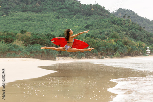 Outdoor photo of gorgeous young woman in fashion bikini doing jump split leap with red surfboard. Summer sunny day sport surfing concept, seascape with girl, beach, beautiful waves, blue water.