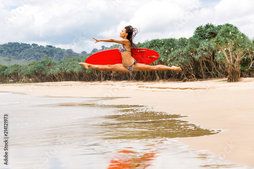 Outdoor photo of gorgeous young lady in fashion bikini doing jump split leap with red surfboard. Summer sunny day sport surfing concept, seascape with girl, beach, beautiful waves, blue water.