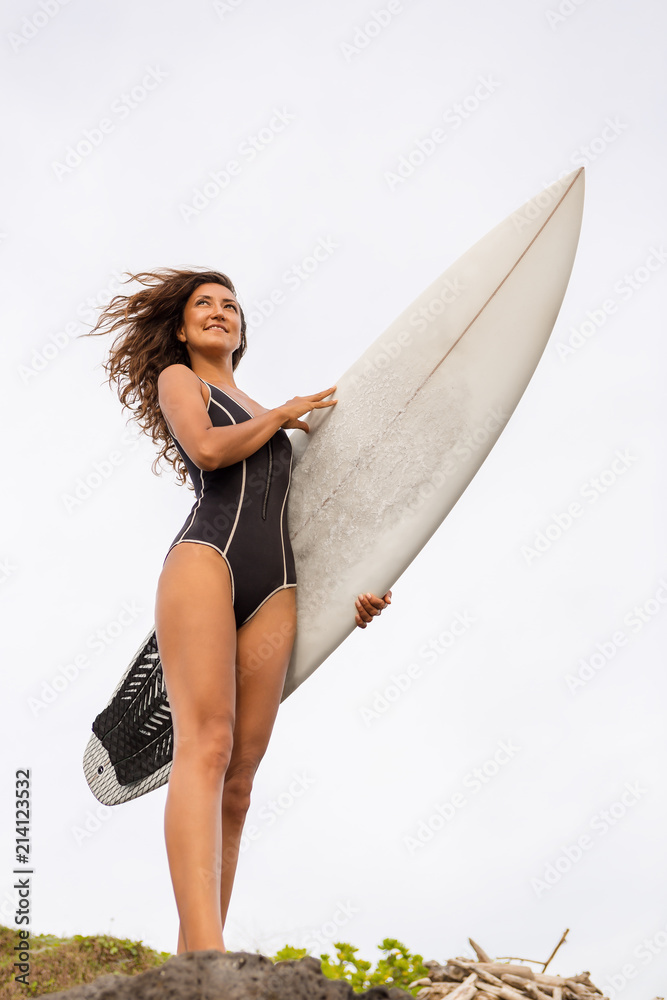 Surf girl with long hair go to surfing. Sporty surfer woman holding  surfboard on a rock