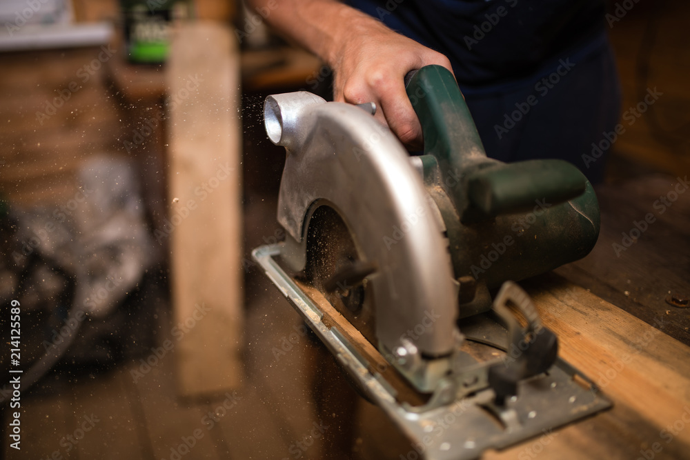 A young male carpenter-carpenter saws a modern circular saw with a wooden board in the workshop, wooden sawdust flying in the sides. to plan a tree. A young male carpenter-carpenter saws a modern circular saw with a wooden board in the workshop, wooden sawdust flying in the sides. to plan a tree.