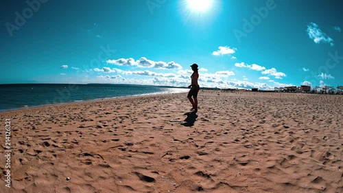 Girl in swimsuit, denim shorts and snapback walking across the beach to water line, summer sunny day at vacation