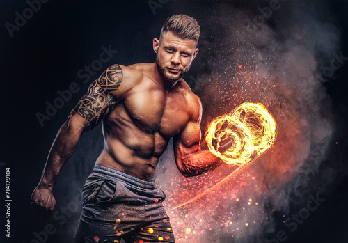 Fototapeta Przystojny wytatuowany kulturysta bez koszuli ze stylową fryzurą i brodą, w sportowych szortach, pozuje w studio. Koncepcja sztuki ognia.