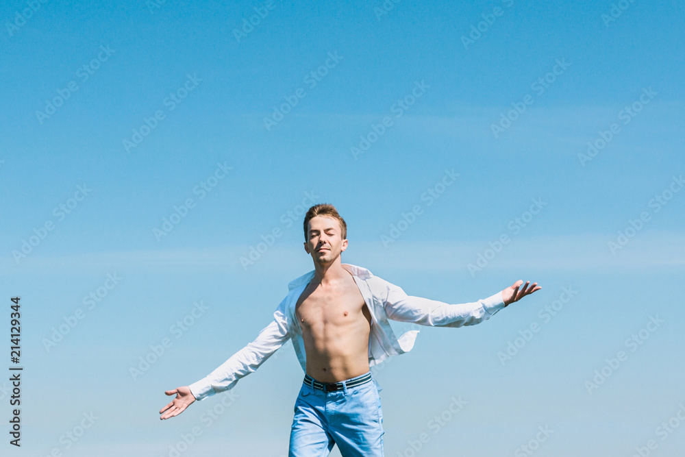 Handsome young man stands with closed eyes against the blue sky with arms outstretched. The white shirt is unbuttoned. Handsome young man stands with closed eyes against the blue sky with arms outstretched. The white shirt is unbuttoned.