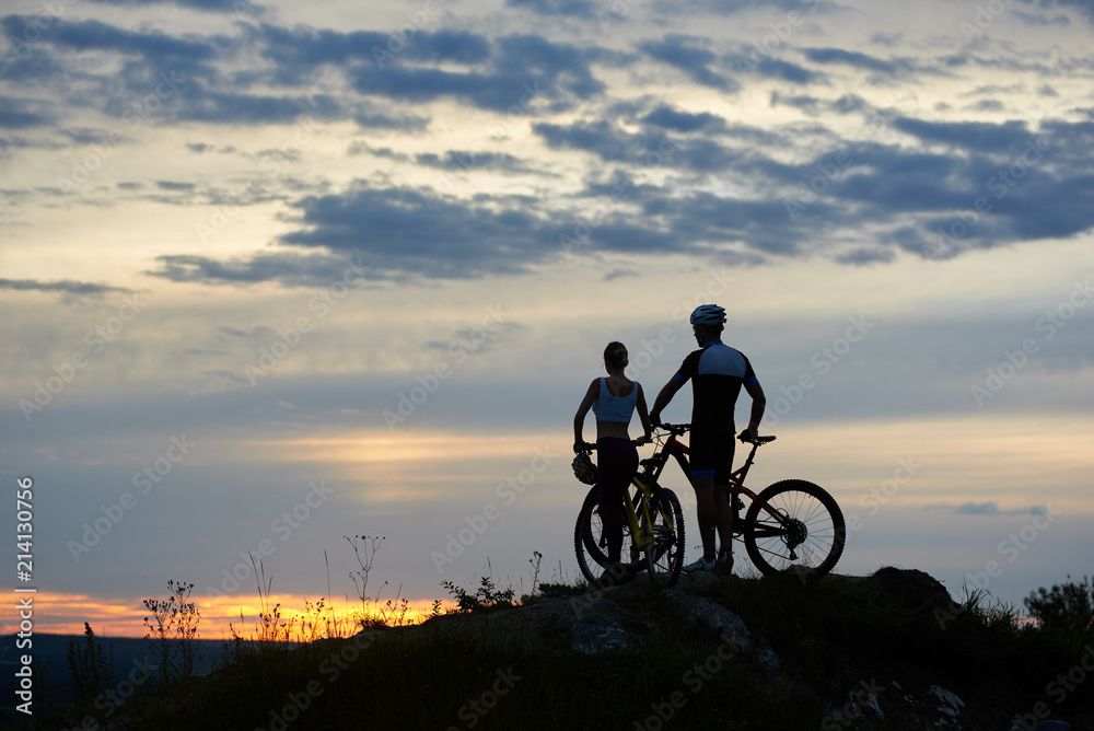 Obraz premium Back view of two young cyclists with mountain bikes standing on top of a cliff with beautiful scenery of hills and sky at sunset. The concept of a healthy lifestyle