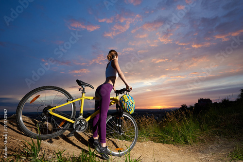 Fototapeta Naklejka Na Ścianę i Meble -  Slender woman wearing violet leggings, posing with her back and buttocks turned to camera. Athletic cyclist standing with yellow bicycle, looking at sunset and beautiful landscape.