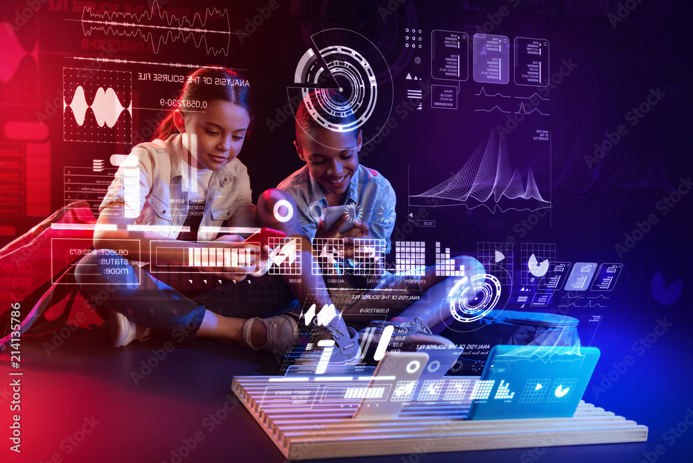 Cheerful friends. Cheerful kids sitting comfortably on the floor in a futuristic room and looking at the modern devices in front of them