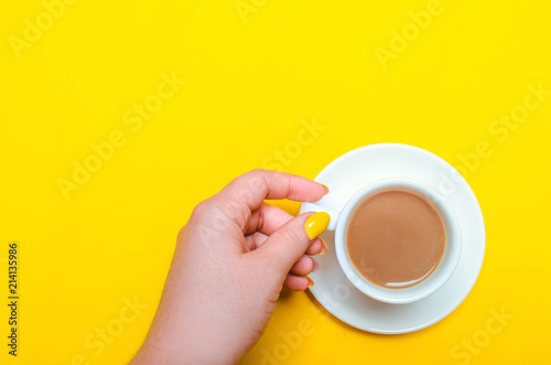 a cup of coffee with a saucer and female hands with a yellow manicure on a yellow background. minimalism. place for text. copy space. top view. flat lay