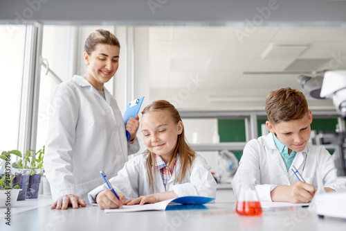 education, science and children concept - teacher and students studying chemistry at school laboratory and writing to workbooks