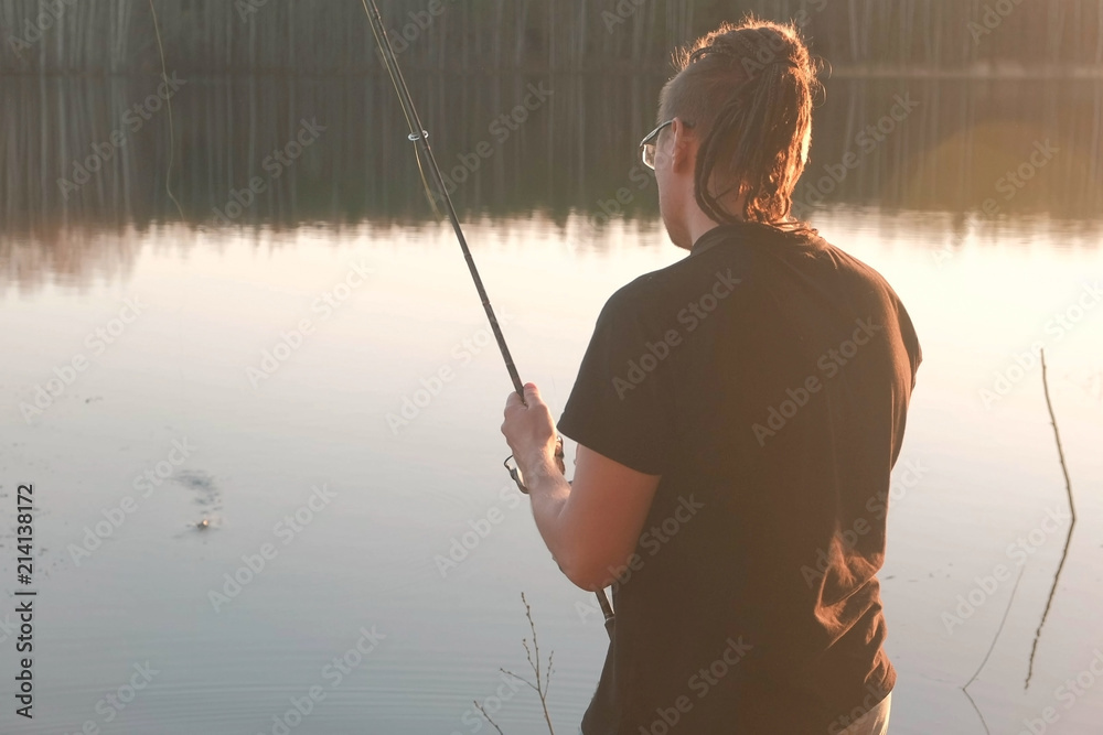 Fisherman on the pond. Young guy with dreads in glasses in a t-shirt  fishing fish with rod. Stock Photo