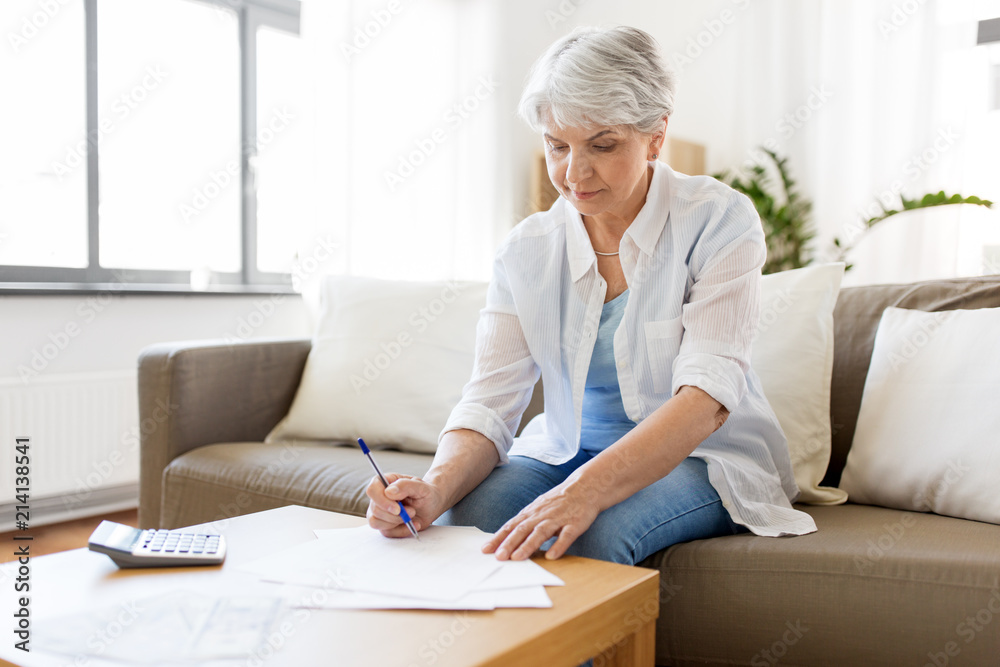 business, savings, annuity insurance, age and people concept - senior woman with papers or bills and calculator writing at home business, savings, annuity insurance, age and people concept - senior woman with papers or bills and calculator writing at home