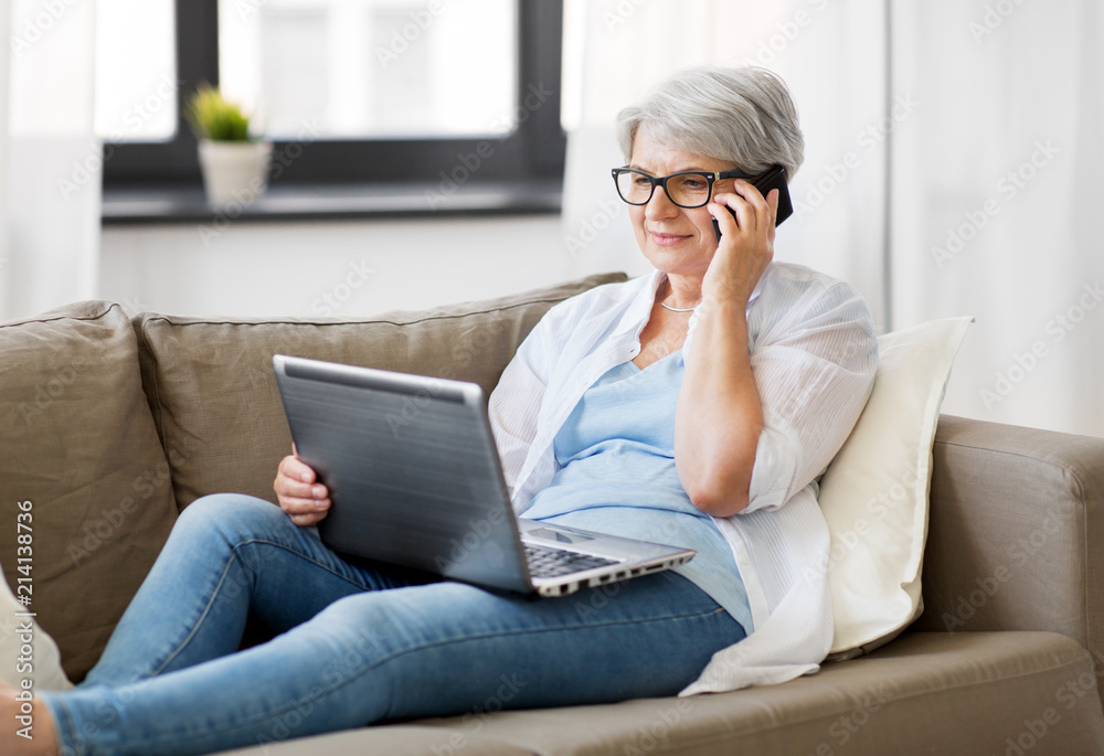 technology, old age and people concept - happy senior woman in glasses with laptop computer calling on smartphone at home technology, old age and people concept - happy senior woman in glasses with laptop computer calling on smartphone at home