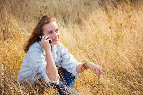 Young smiling blond girl sitting in golden field and talking on phone.