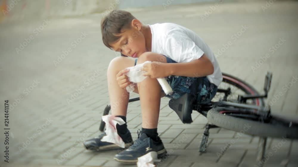A young boy sits in a park after falling from a bike, calms the pain in his knee, is a dangerous bike ride A young boy sits in a park after falling from a bike, calms the pain in his knee, is a dangerous bike ride