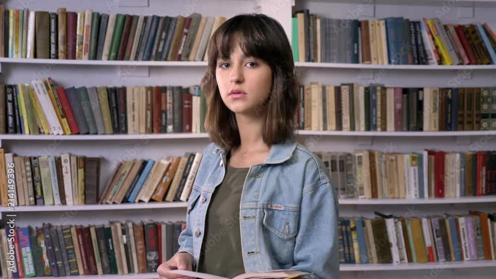 Young brunette girl is typing reading book, watching at camera, library on background Young brunette girl is typing reading book, watching at camera, library on background