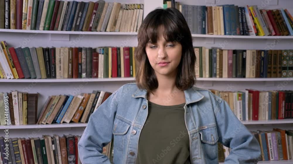 Young brunette girl is standing with hands on hips, watching at camera, library on background Young brunette girl is standing with hands on hips, watching at camera, library on background