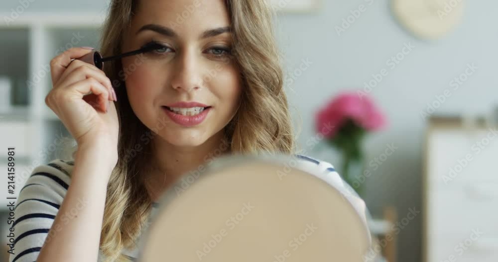 Portrait shot of the charming young Caucasian woman painting her eyelashes in front of the mirror. Beauty blog. Indoors. Portrait shot of the charming young Caucasian woman painting her eyelashes in front of the mirror. Beauty blog. Indoors.