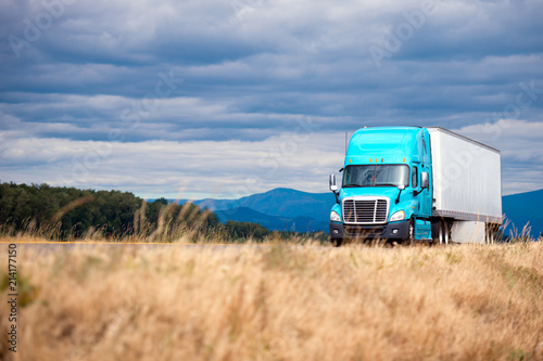 Blue modern big rig semi truck transport semi trailer on scenic road with grass forest mountain and cloudy sky