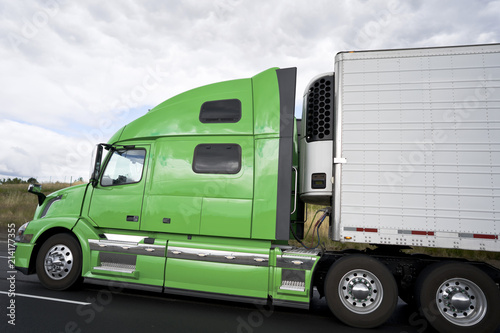 Profile of green big rig semi truck with refrigerated semi trailer on the road
