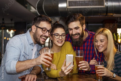 Goup of four friend in local pub looking at mobile phone and smile with glass of beer on hands