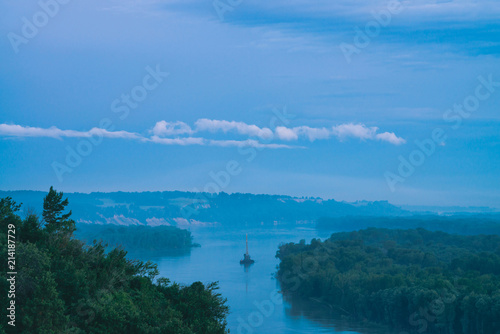 Beautiful river night landscape with green shores and ship with copy space. Towboat tows barge with crane along riverbank at dusk. Blue water in evening. Calm time. White cloud in sky on background.