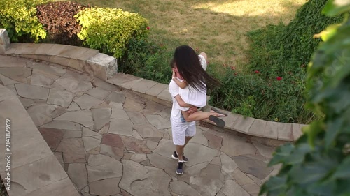 The happy guy holds his girlfriend in his arms and turns around, the girl enjoys and waves her long hair. Beautiful loving couple walking in the summer park. Slow motion. photo