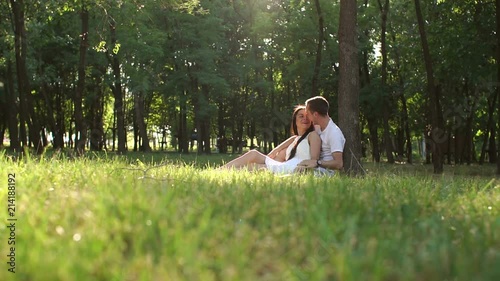 Young teen lovers are sitting on the grass leaning against a tree in the Park in Sunny warm weather. Wide shot. Slow motion. photo