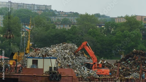 In the seaport of the city of Vladivostok, the excavator is unloading scrap-metal from the barge at the pieron the background of urban high-rise buildings photo