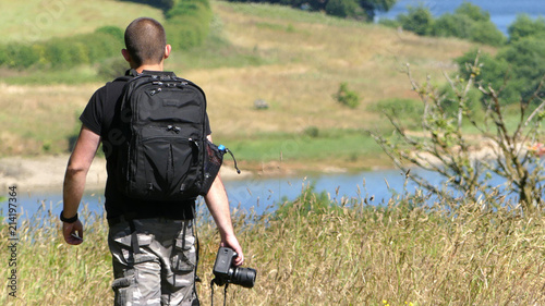 A male photographer sets off towards a lake, wearing shorts and rucksack, camera at his side